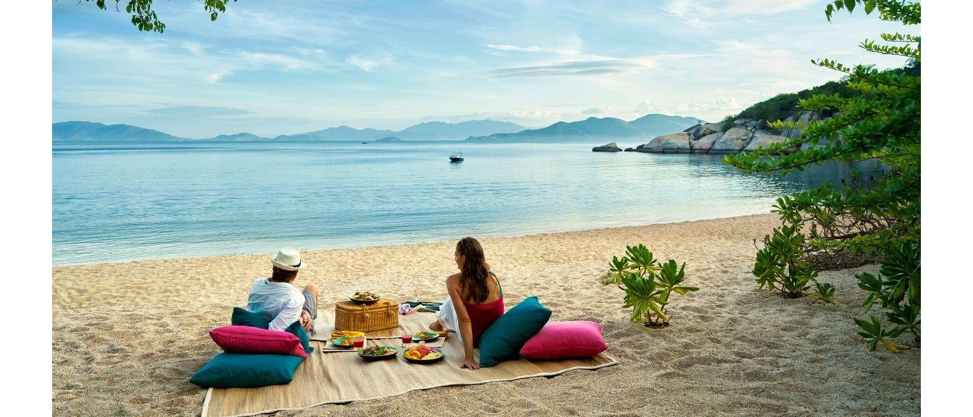 Couple gaze out to the sea as they enjoy a beach picnic on pink and blue cushions, with a blanket, picnic basket and plates of salad and fruit between them