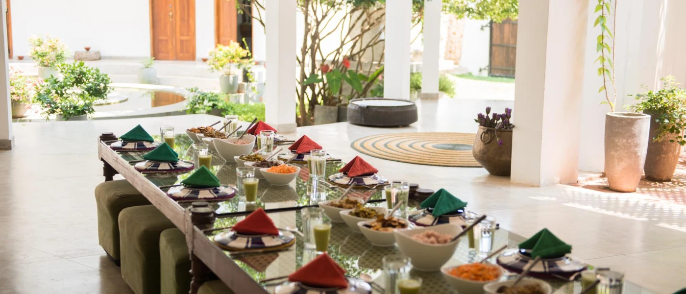 Communal table laid up for a meal, with a line of white bowls filled with food, and each plate next to a glass with green juice inside