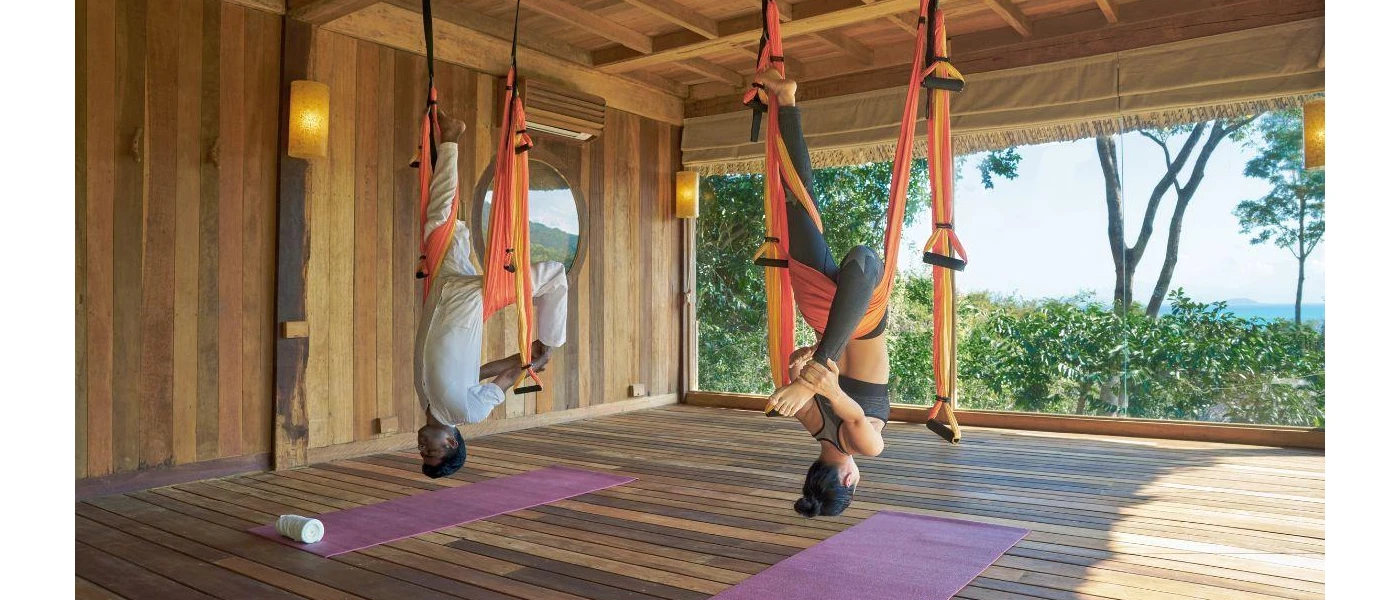 Man and woman hang from the ceiling in acrobatic ribbons, in a wooden studio space with a large window facing the sea