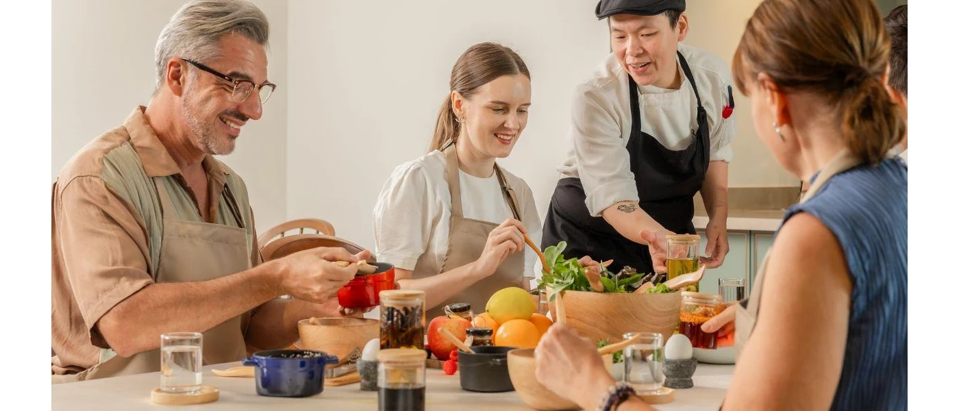 Family in aprons sit around a table spooning things out of jars, as a chef watches on