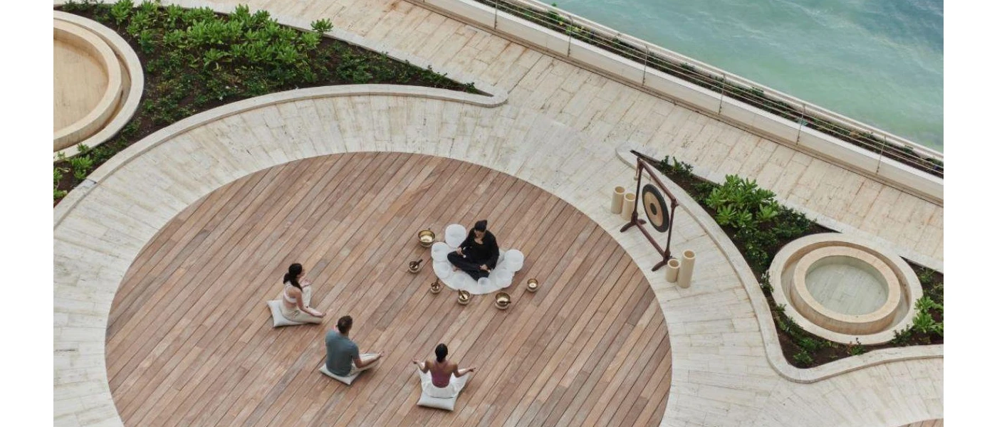 Three figures seen from above, sitting cross legged on a round wooden deck as an instructor surrounded by sound bowls and with a gong behind her, takes a healing class