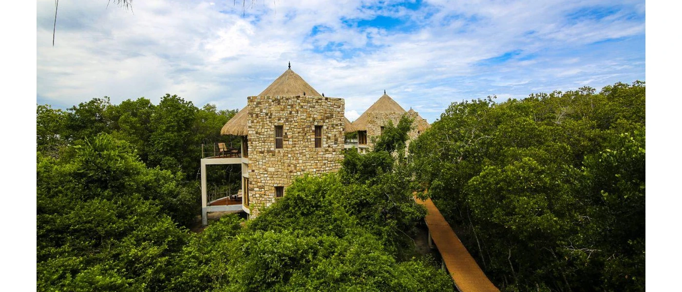 Stone buildings with thatched roofs next to a forest and a footbridge