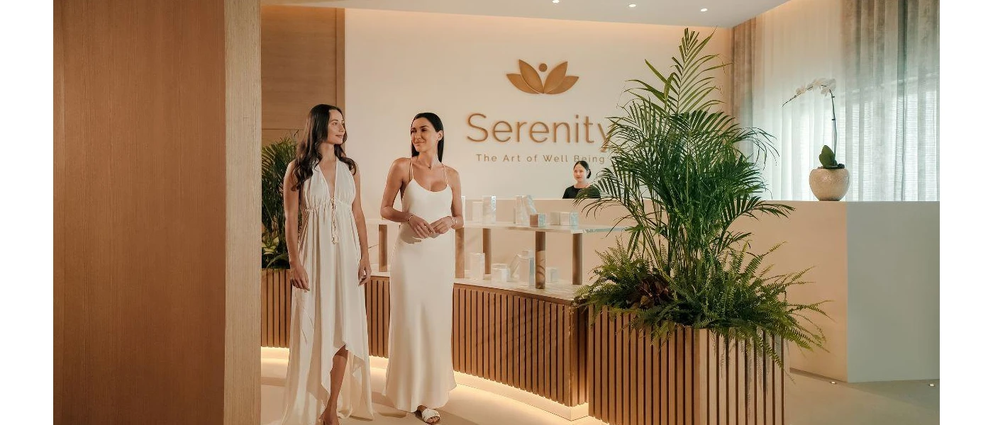 Smiling women in white dresses stroll through a reception area with wood-panelled walls, soft lighting and plant beds, with a sign that reads 'Serenity the art of wellbeing' behind them