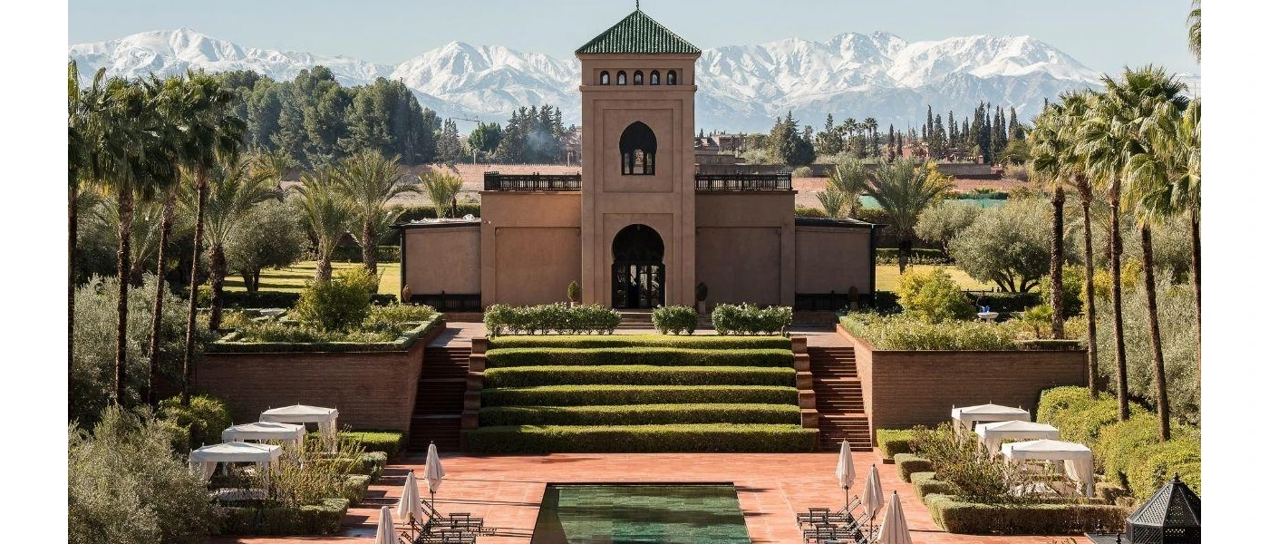 Grassy staircase in front of a decadent building with tower, rectangular swimming pool and daybeds with white curtains