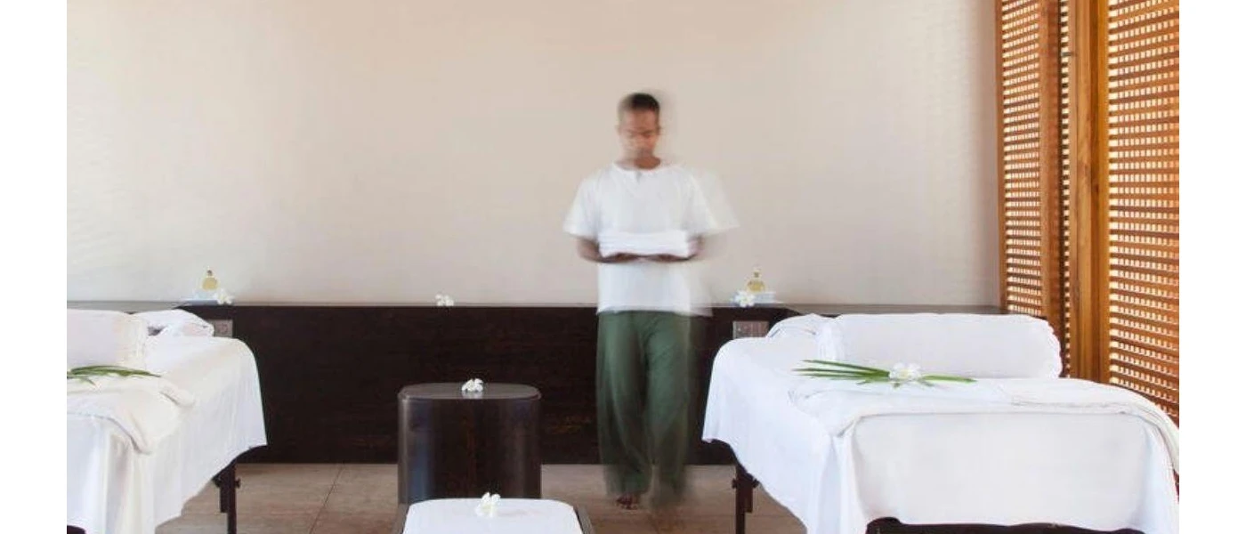 Man in airy clothes holding a tray in a spa treatment room with two massage beds covered in white cloth