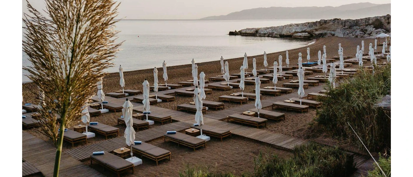 Sandy beach with wooden walkways, cushioned loungers and white parasols, looking out onto a curved bay under a cloudy sky