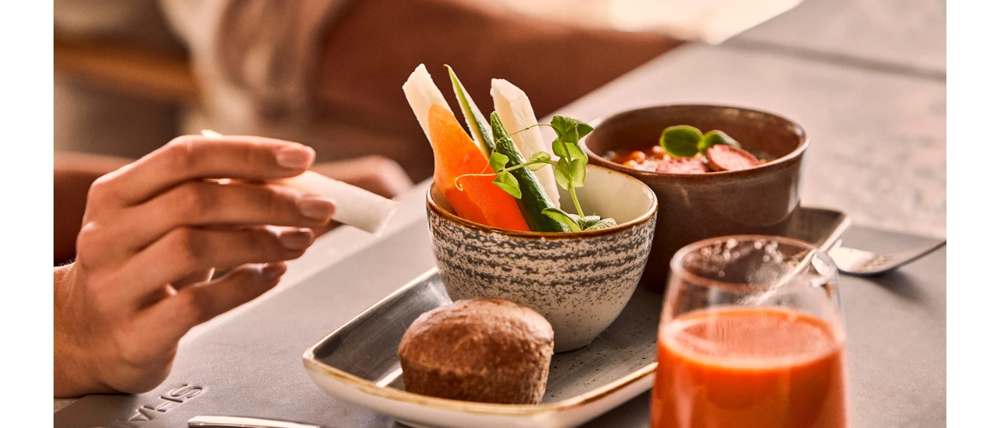 A bowl of vegetables and a bread roll on a tray, next to a juice or smoothie with an orange colour