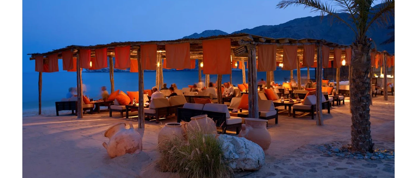 Open-air bar on the beach, with orange material draped around the sides, and low-rise tables and chairs, as the sky darkens