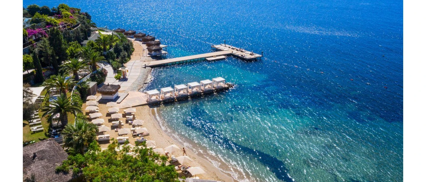 Palm-lined lawned terraces leading down to a sand and shingle beach overlooking jetties and turquoise waves
