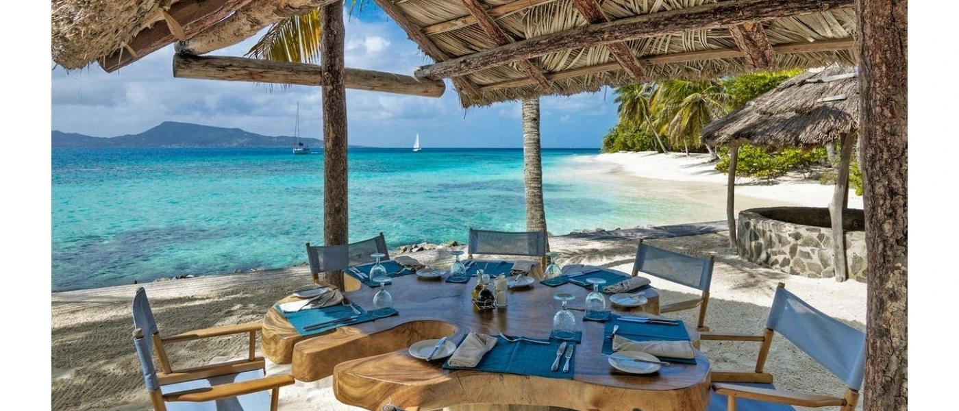 Thick wooden table set up with blue napkins and place mats under a thatched roof overlooking the ocean