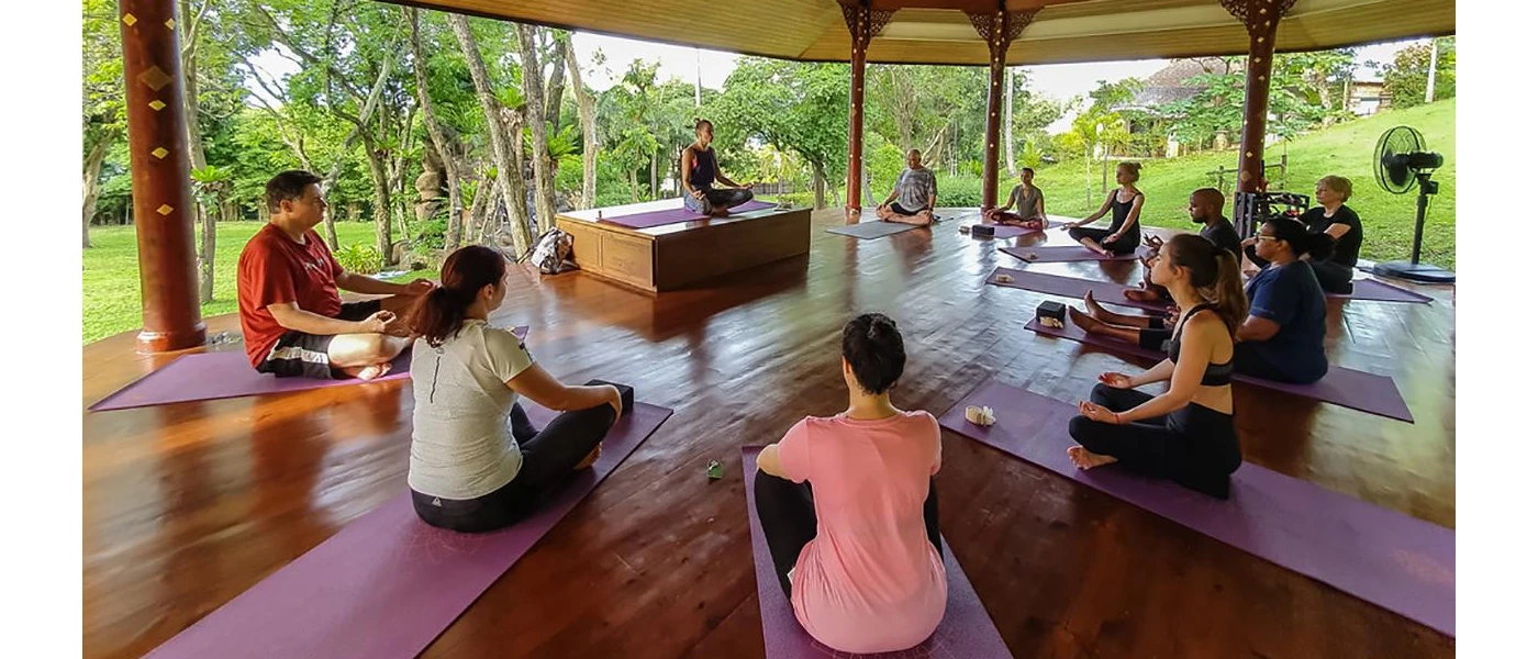 Group sit cross legged on mats in a circle in a open-sided yoga pavilion, as an instructor demonstrates on a raised block upfront 