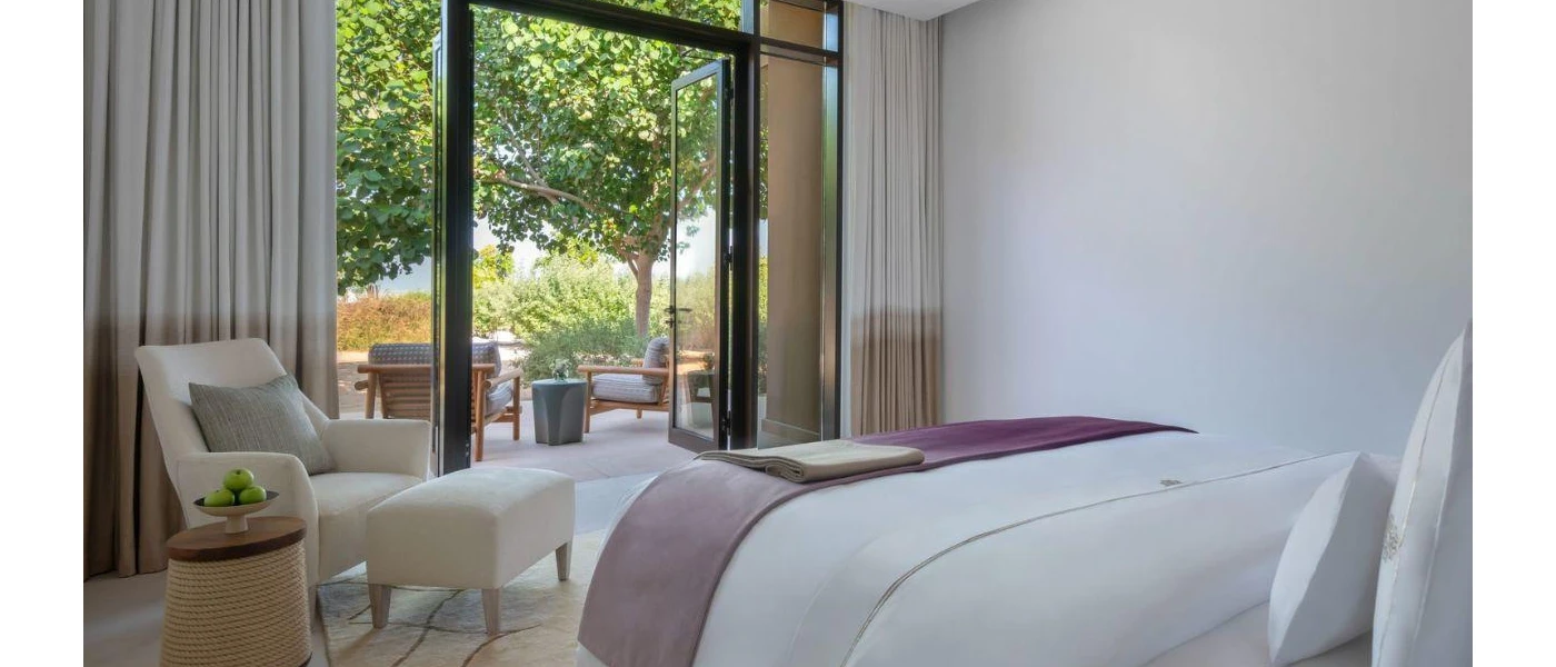 Hotel bedroom with white bedding and a grey throw, a white tub chair and footstall in one corner and a stall with a bowl of apples, with floor-to-ceiling balcony doors in front