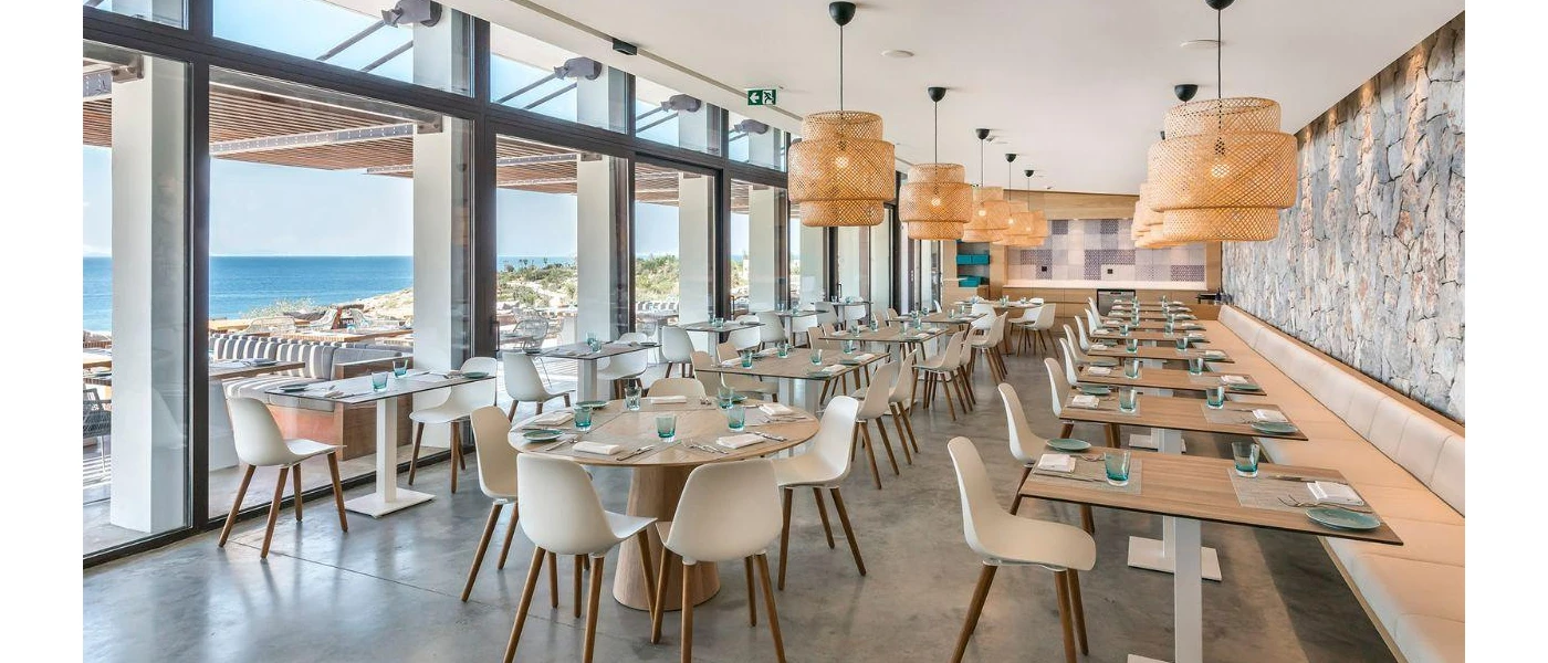 Bright restaurant with floor-to-ceiling patio doors, rattan hanging pendant lights, simple wooden tables and a sea view