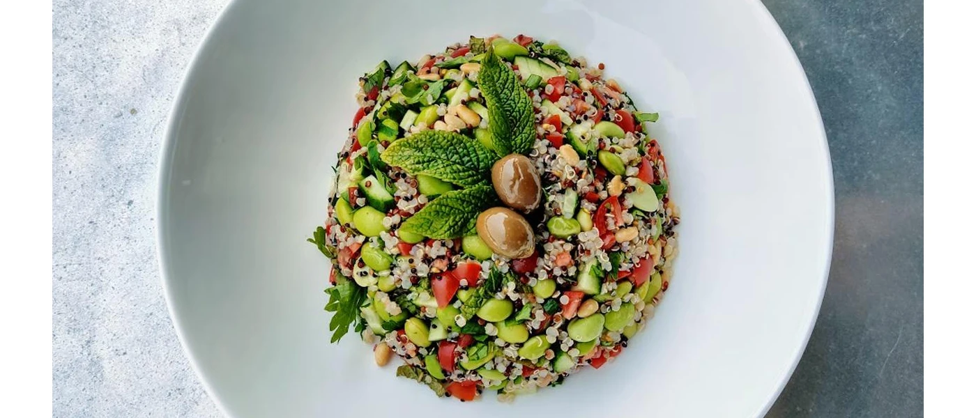 White bowl with grains, tomatoes, edamame beans and cucumber, with a mint and olive garnish