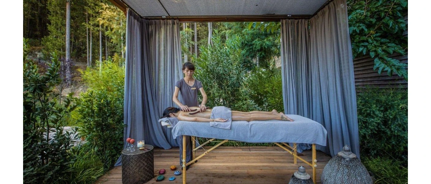 Woman lies partially covered by a blue towel on a massage bed in an open-air cabana surrounded by a forest, as a therapist massages her back