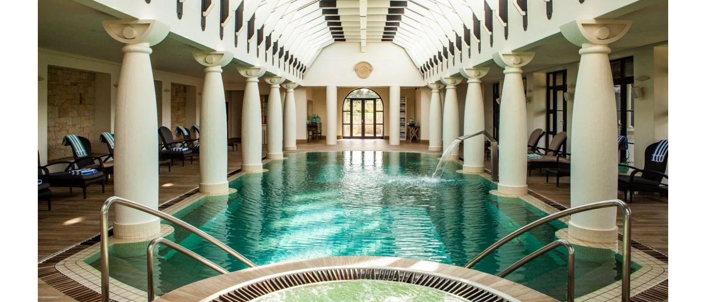 Indoor pool with a whirlpool bath section, in a grand room with white pillars and dark loungers with blue and white striped towels