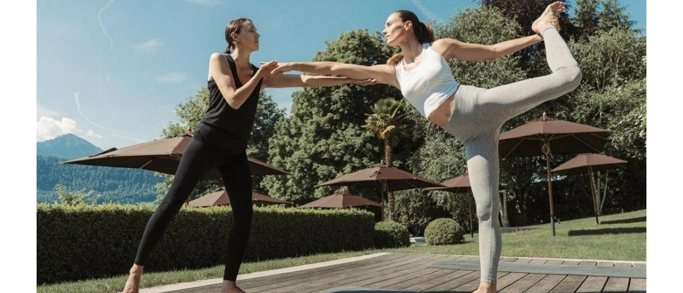 Woman in active wear in a yoga pose, being guided by an expert in a sunny, leafy outdoor garden