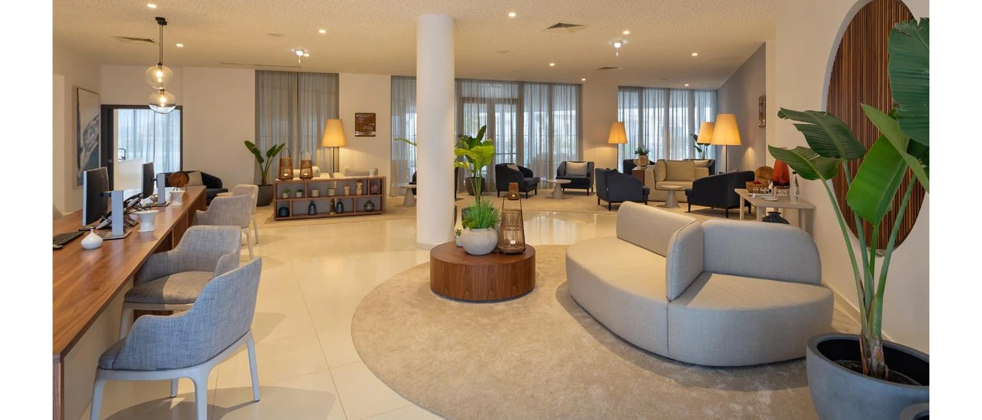 Lobby lounge area with ceiling spotlights, white pillars and walls, a glossy white floor, grey sofas and chairs, potted plants an dark-wood desks