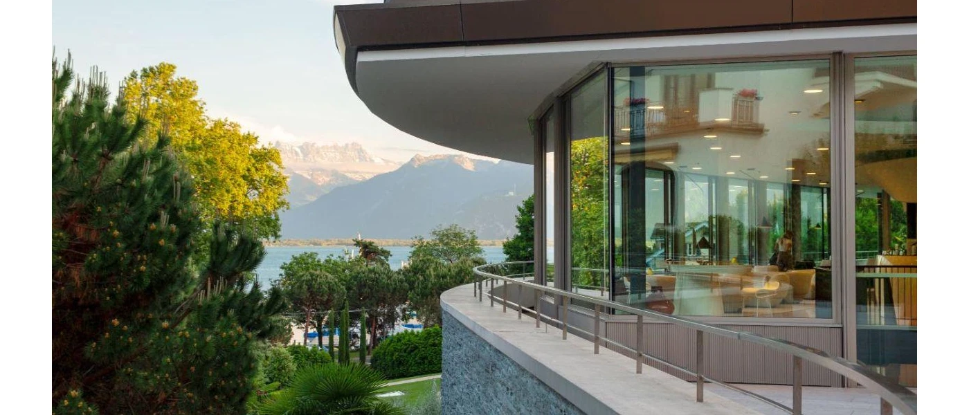 Restaurant with floor-to-ceiling windows and a wraparound balcony overlooking gardens, a lake and mountains