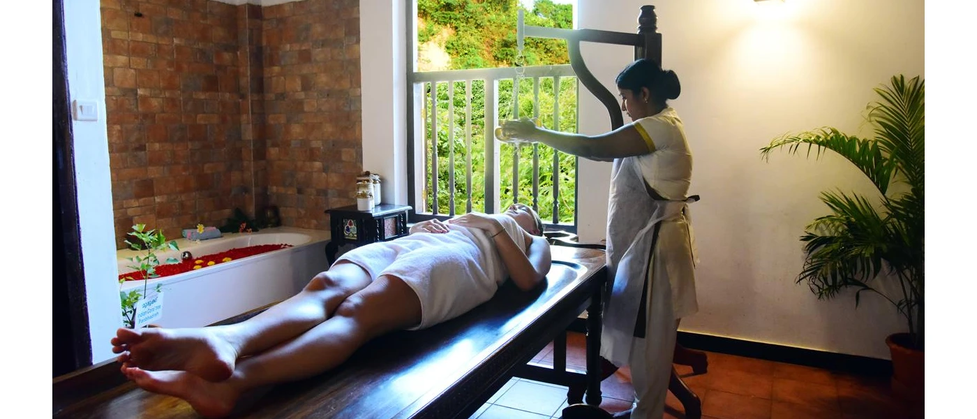 Woman lies on her back in a white towel on a bed in a treatment room with white walls, a full-length window and bathtub filled with red rose petals, as a woman prepares to pour oil over her head