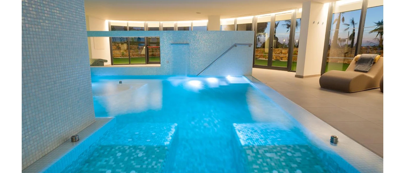 Indoor pool with tiled floors and surrounding walls, floor-to-ceiling windows to the right-hand side and contemporary cream squishy chairs