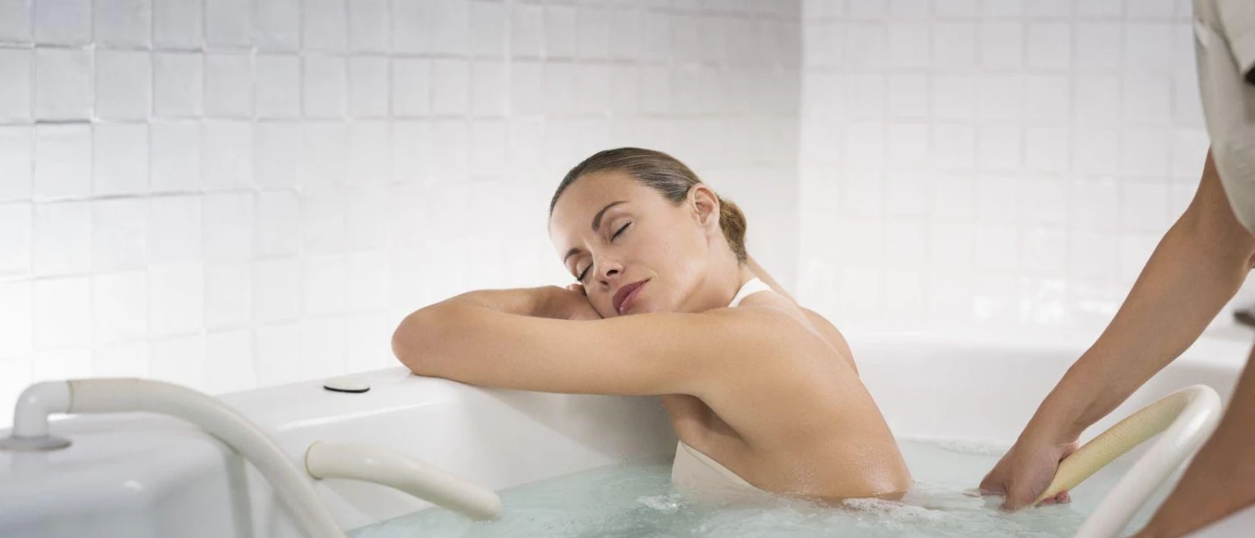 Woman rests with her eyes closed and her head on her arms in a bath as a therapist tests the temperature