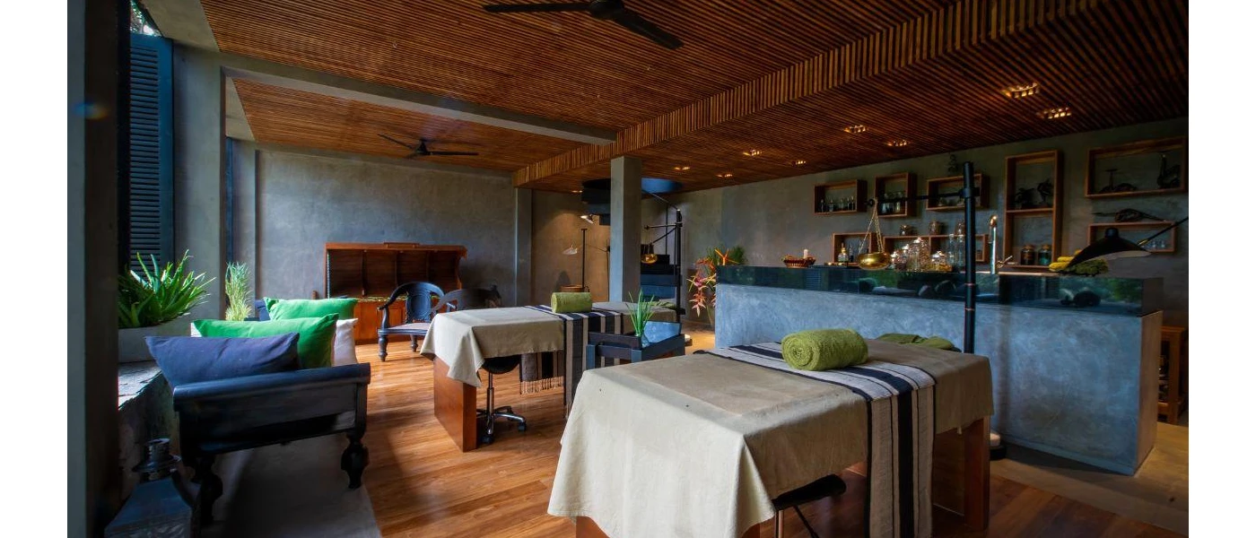 Treatment room with two massage beds, a wall filled with shelves lined with lotions and potions, and a wooden ceiling with black fans
