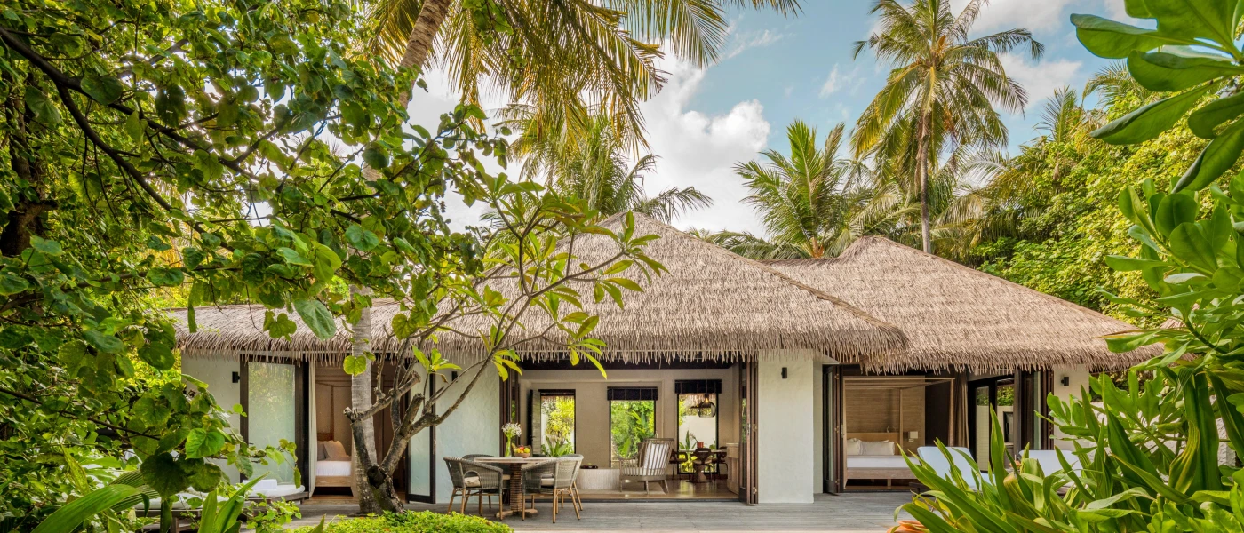 Thatched-roof villa surrounded by palm trees and shrubs, with a wooden deck furnished with a table and chairs