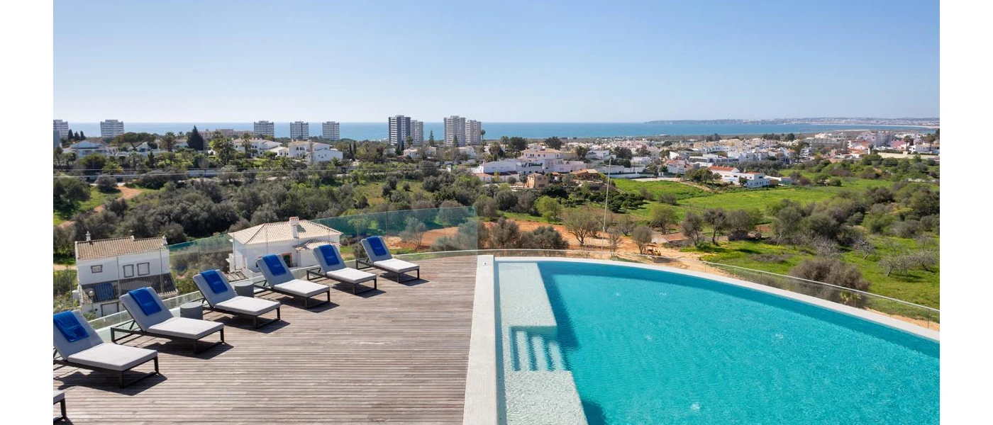 Rooftop with wooden decking and a swimming pool, with ivory loungers and a glass balcony with views of surrounding greenery and a whitewashed town
