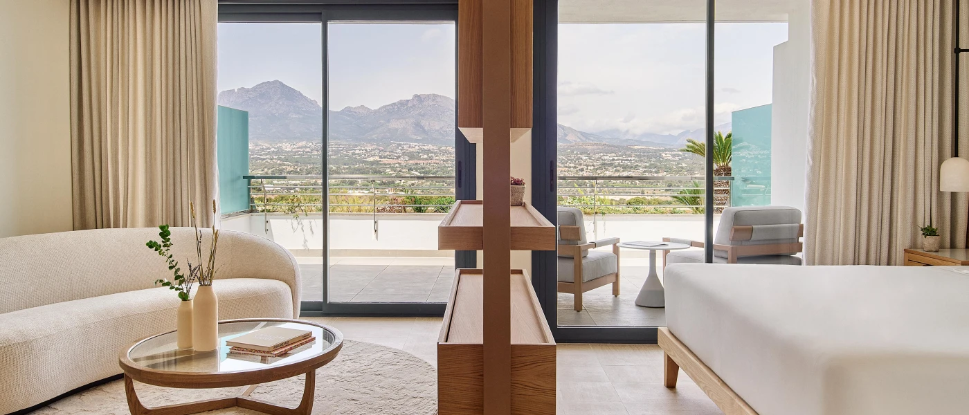 Bright hotel room with white bedding, a cream sofa, glass-topped coffee table and wooden unit, plus floor-to-ceiling windows showcasing a furnished balcony and views of the mountains