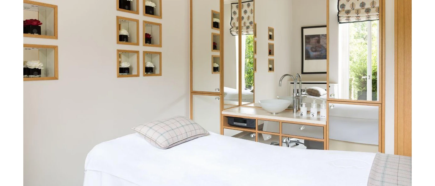 White treatment room with wooden shelves fitted with pots of flowers and a vanity unit with a standalone sink next to a bed