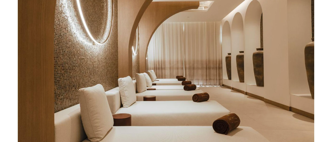 Softly lit relaxation room with a line of white-cushioned beds, a tiled wall, archways decked out with oversized vases and brown rolled up towels on each bed