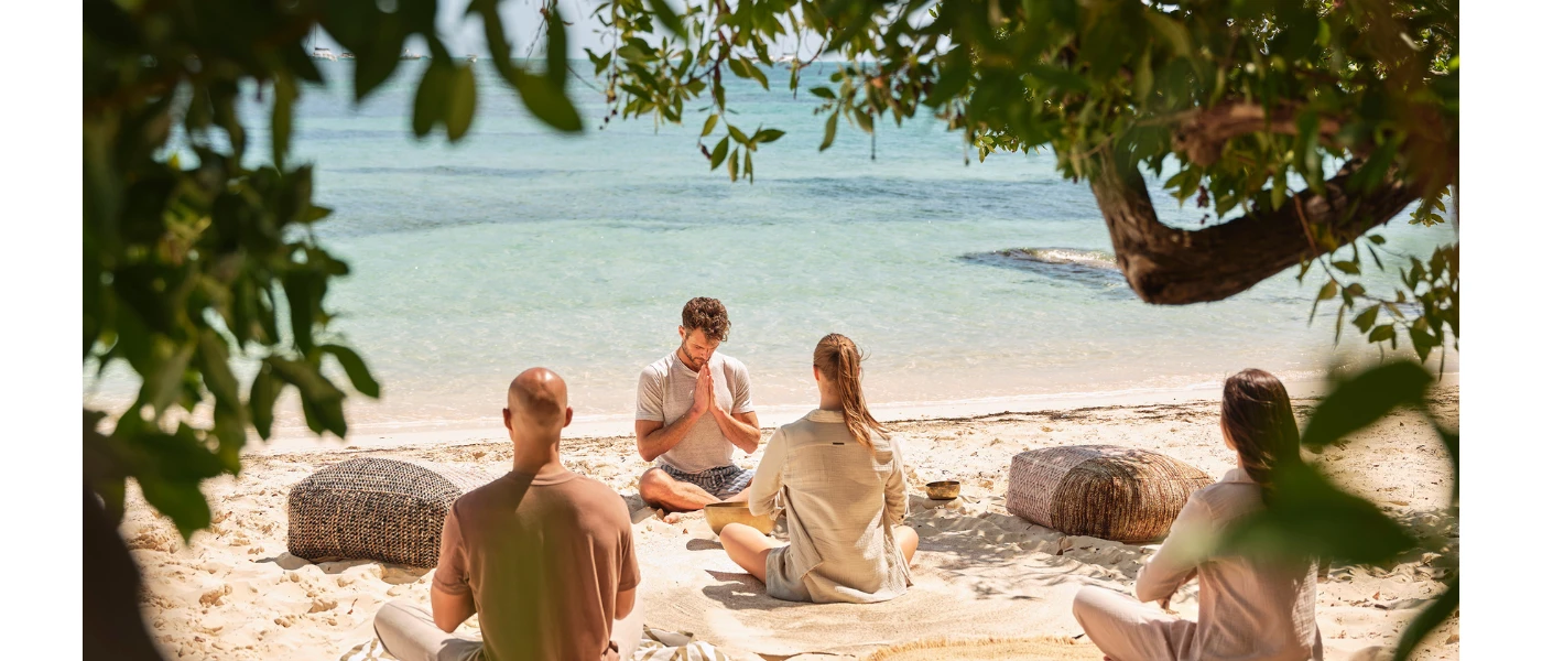 Three people sitting cross legged with their hands in prayer on the beachfront, as an instructor leads from the front