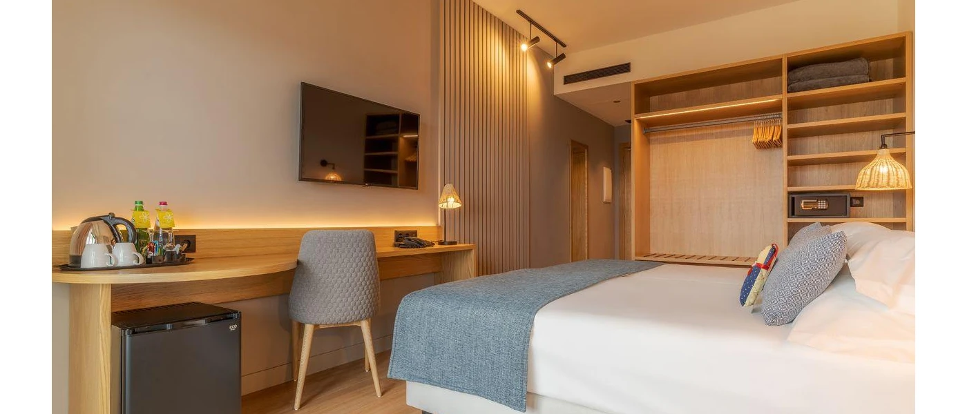 Hotel room with a light-wood wardrobe and desk, white walls and a white bed with blue soft furnishings