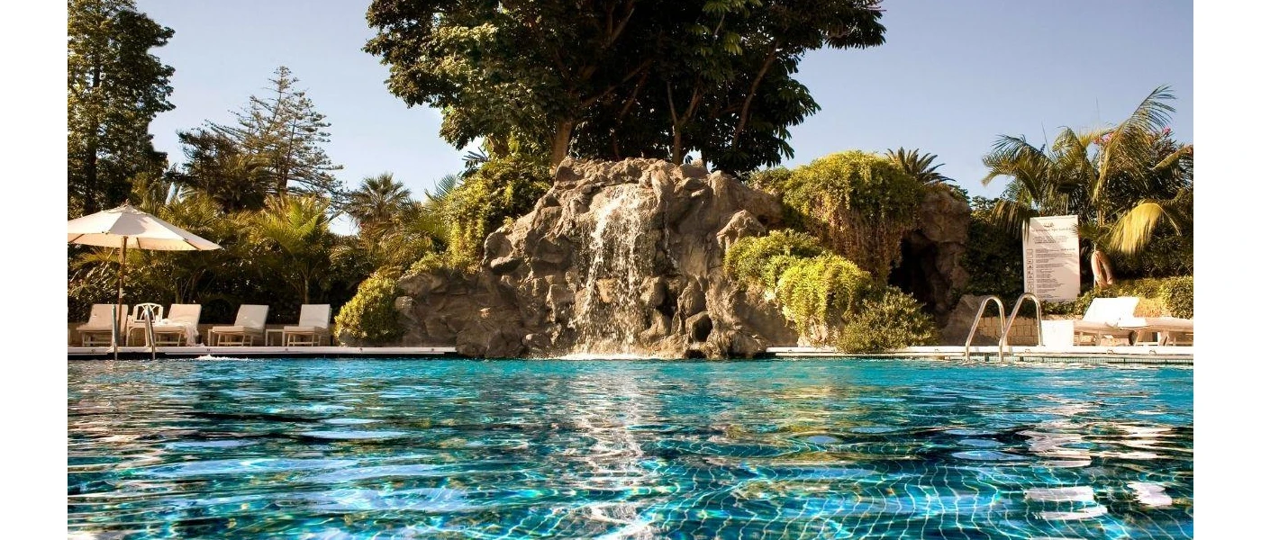 Huge pool with rockery and a waterfall in the middle, surrounded by tropical gardens, white parasols and loungers