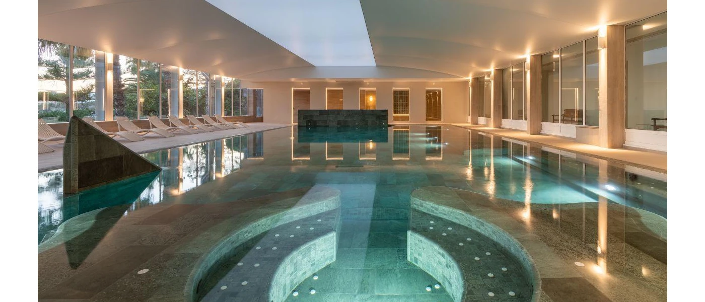 Whirlpool section in an indoor pool, surrounded by white loungers and full-length windows with gardens one side, and seating areas the other