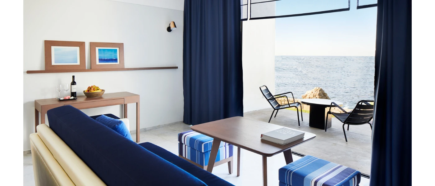 Modern hotel room with blue and white seating, a wooden coffee table and furnished balcony overlooking the sea