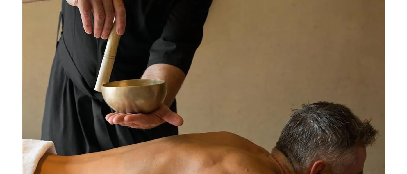 Man lies on his front in a white towel, as a therapist in black uniform carries out sound healing over the top of him