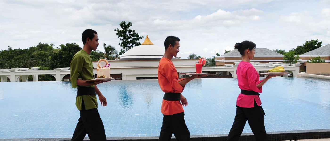 Three staff members in pink, orange and green uniforms carrying colourful drinks past a large pool