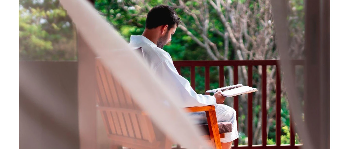 Man in white reads in a wooden chair on a balcony