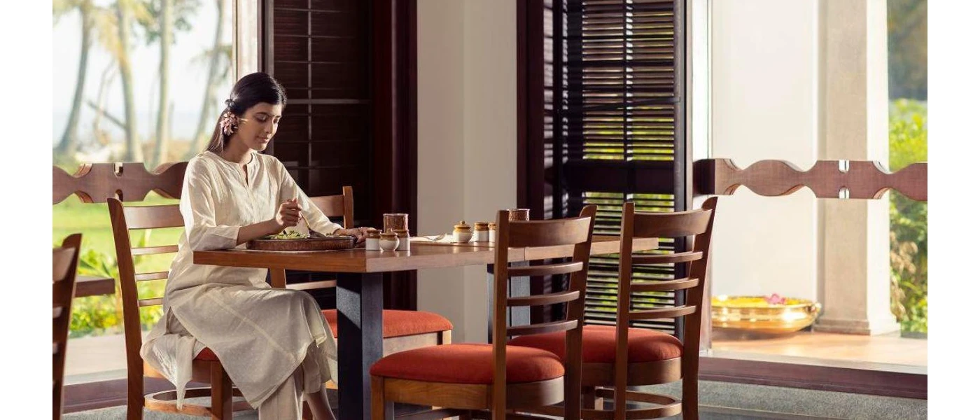 Woman in white with a pink flower in her hair sits eating at a wooden table in an airy restaurant