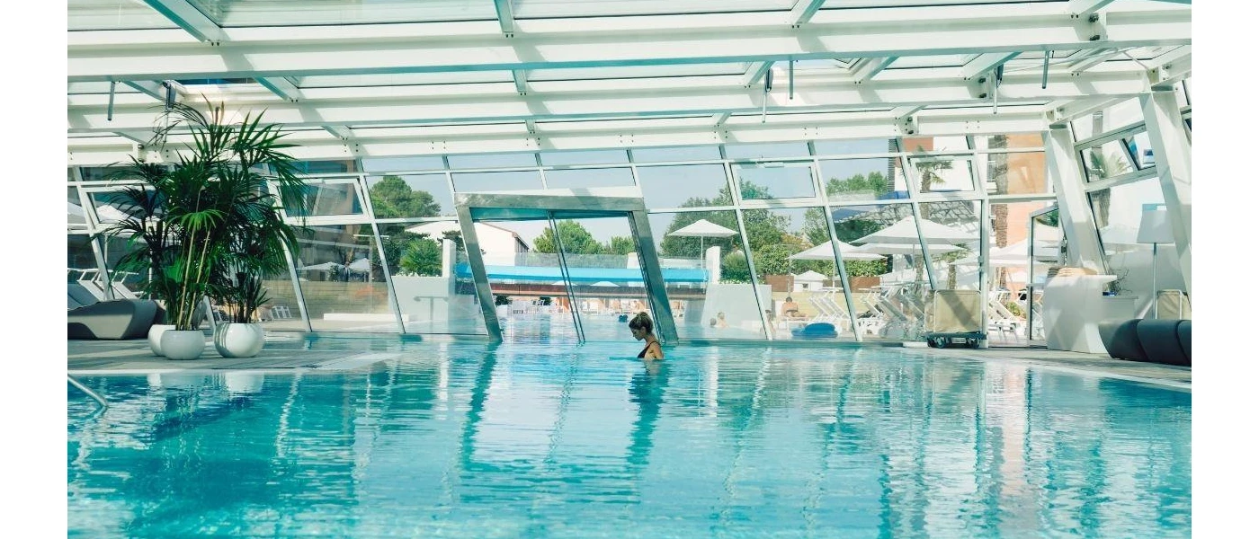 Woman in a black swimsuit relax in an indoor pool that leads outdoors with wraparound glass windows and ceiling