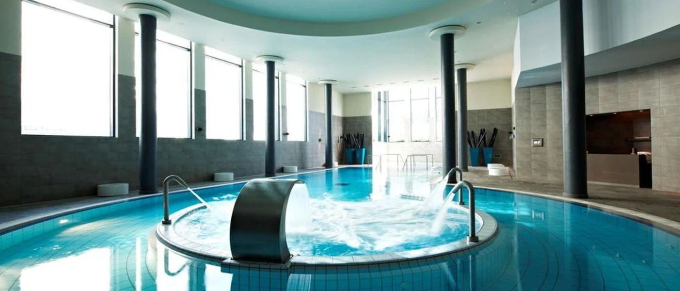 Circular indoor pool with a raised whirlpool section at the centre, surrounding dark pillars and large windows