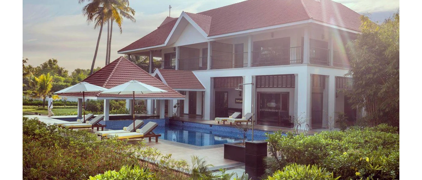 Rectangular swimming pool surrounded by white-cushioned loungers and parasols, in front of a white two-storey building with a terracotta-tiled rooftop