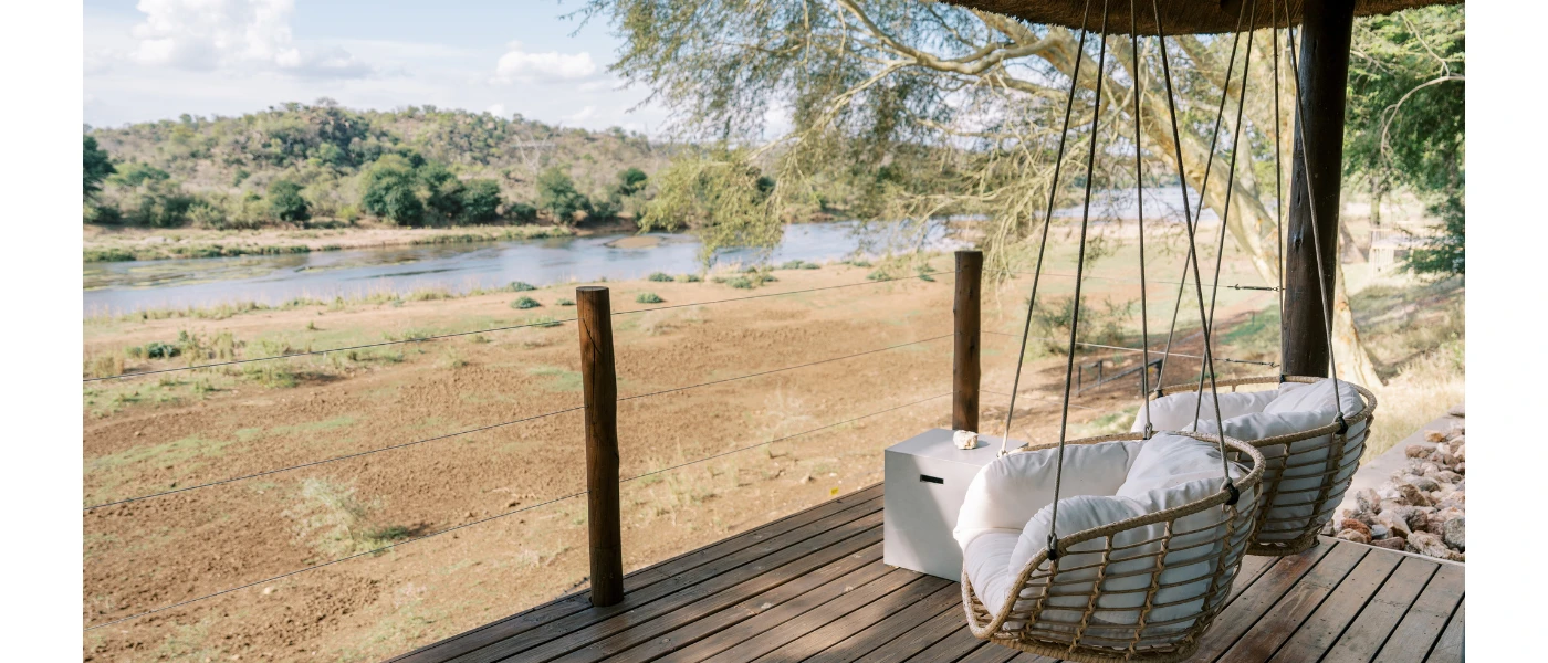 Wooden deck with hanging rattan chairs and white cushions, overlooking the river bank and water