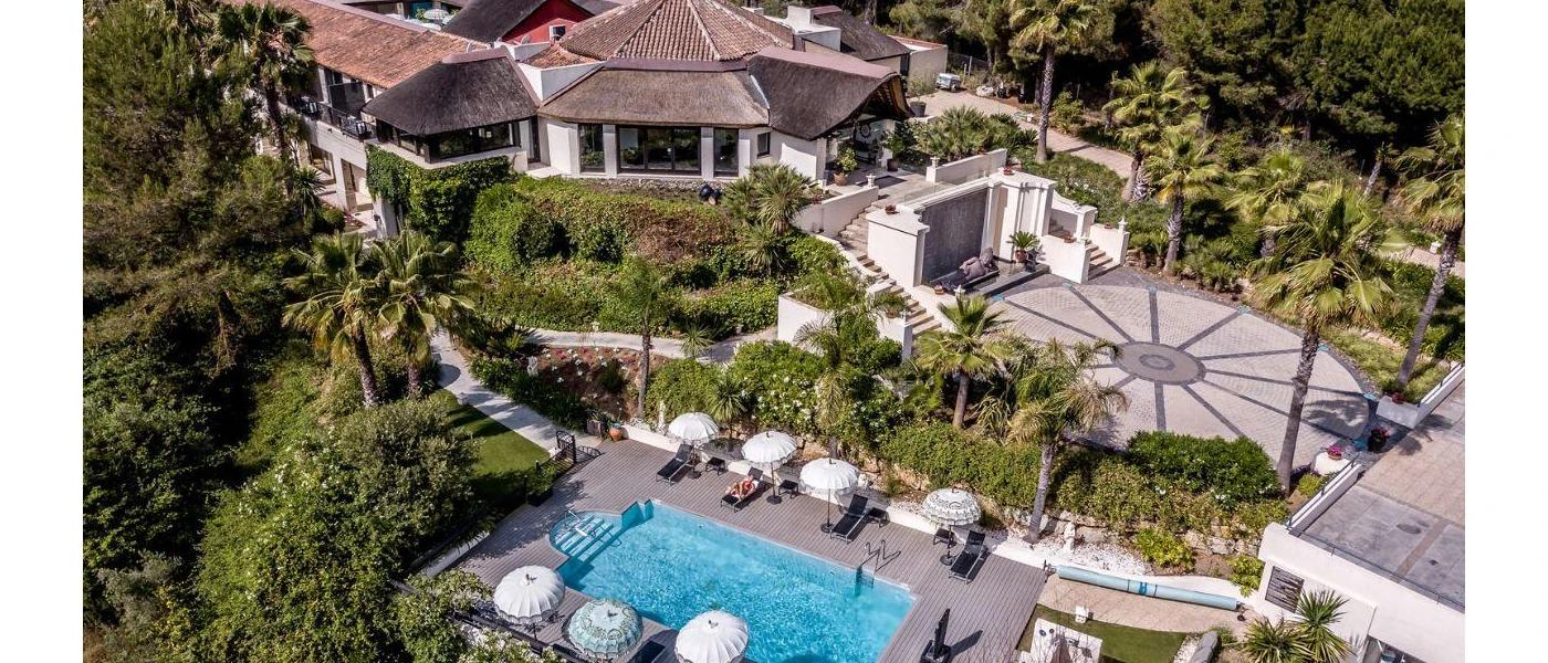 Low-rise buildings with grey rooftops, tropical gardens, a landscaped driveway and a pool lined with white loungers, as seen from above