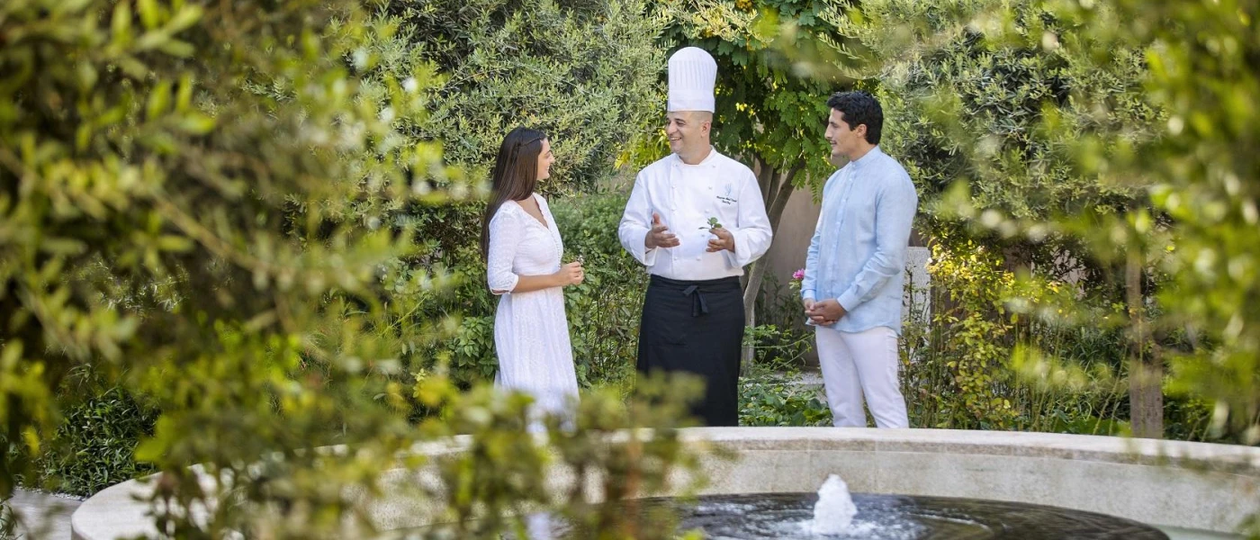 Chef in hat and apron chats with a couple in a garden courtyard with a water fountain