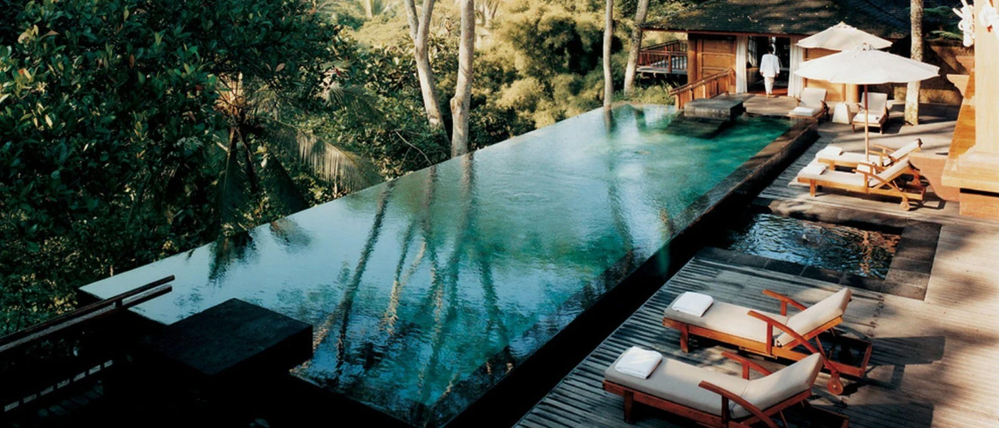 Infinity pool overlooking tropical greenery, edged with wooden loungers with white cushions, white umbrellas and stone steps into the water