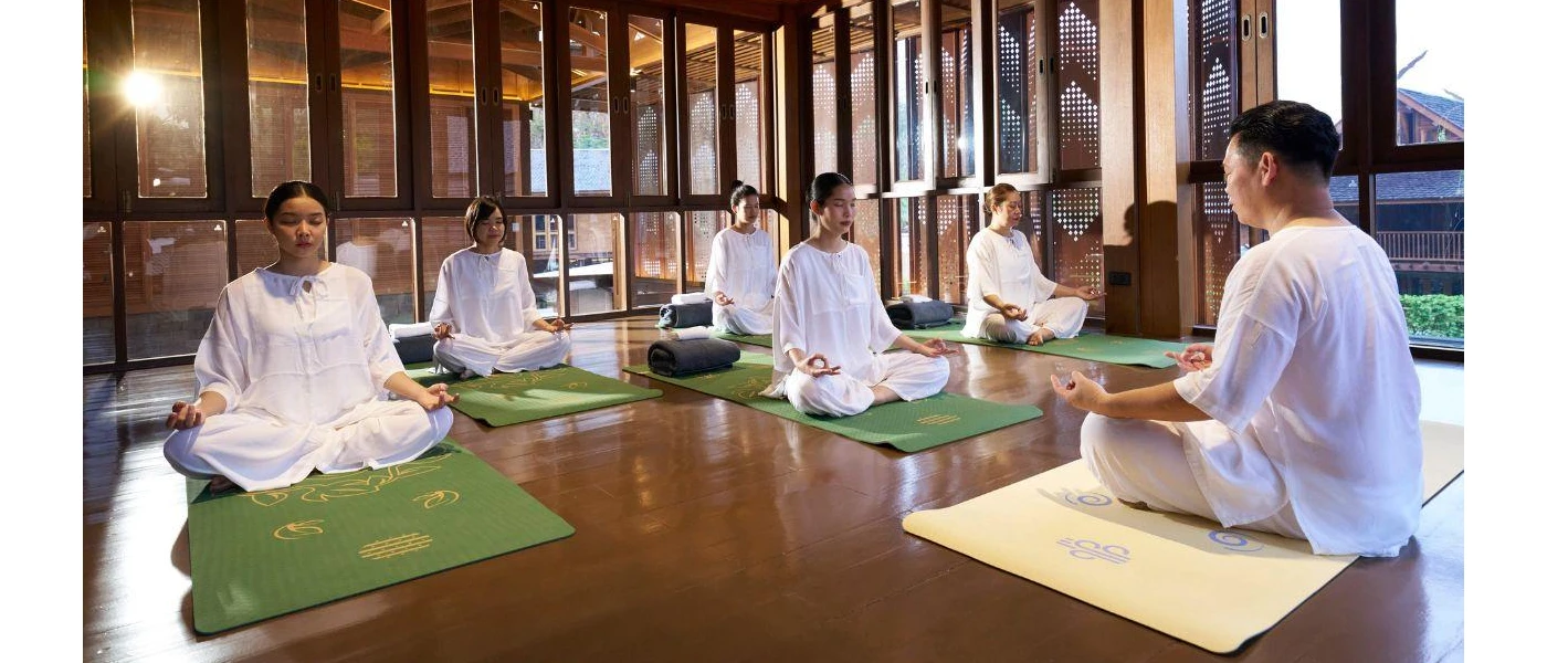 Group in white on green mats with closed eyes, legs crosses and their palms upwards, in a wood-walled, wooden floored studio