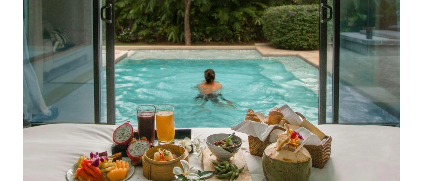 Man swims in a pool in front of a bedroom laid up for breakfast with fresh fruit, coconut juice, a basket of pastries and fresh juices